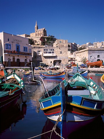 Fishing boats luzzu in Mgarr harbour with Our Lady   of Lourdes Church on the hill     Gozo Malta
