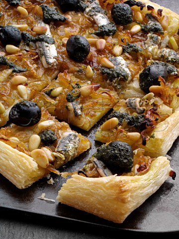 Pissaladire pizza with black olives anchovies and pine nuts on black baking tray