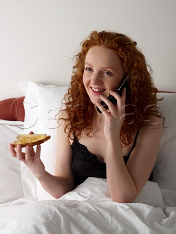 Young woman eating toast and marmalade in bed whilst talking on her mobile phone