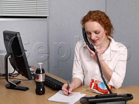 Young woman making notes whilst on telephone at her office desk bottle of diet coke and bag of lofat crisps