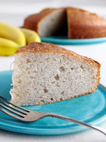 A slice of banana cake with a whole cake and fruit behind
